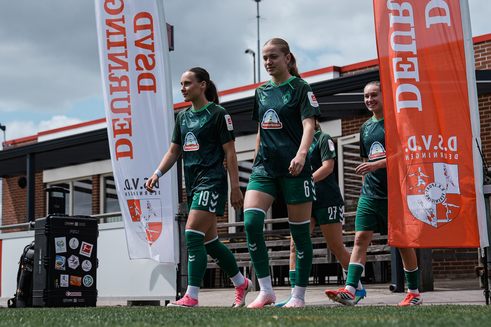 Fußballspielerinnen in grünen Trikots betreten unter bewölktem Himmel das von orangefarbenen Bannern gesäumte Spielfeld.