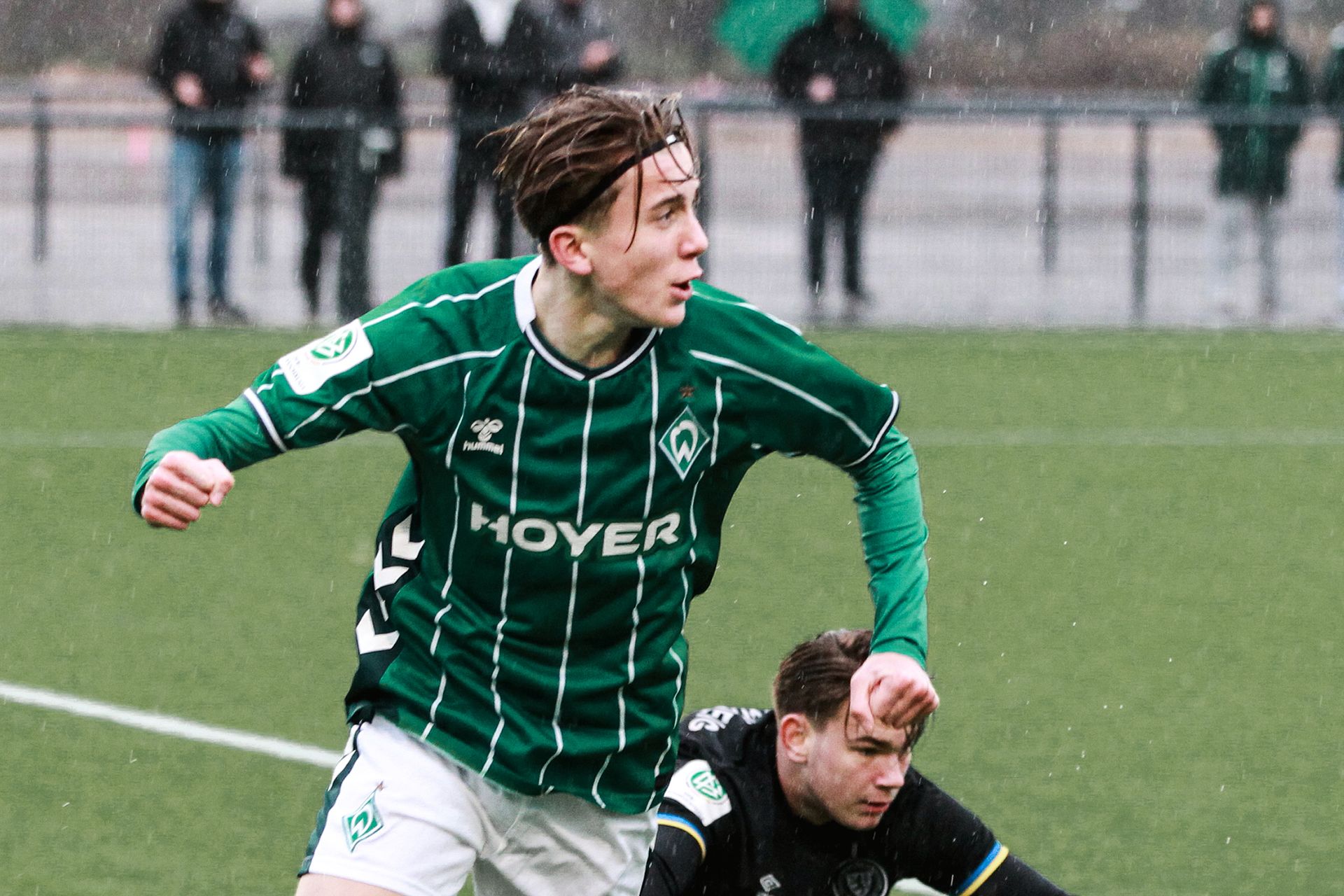 Ein Fußballspieler in einem grün-weiß gestreiften Trikot rennt über einen regennassen Platz, hinter ihm liegt ein anderer Spieler auf dem Boden.