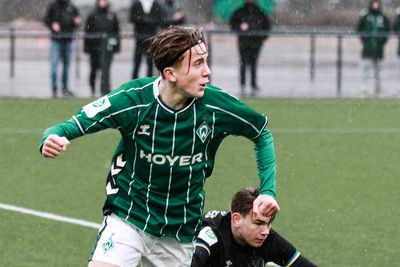 Ein Fußballspieler in einem grün-weiß gestreiften Trikot rennt über einen regennassen Platz, hinter ihm liegt ein anderer Spieler auf dem Boden.