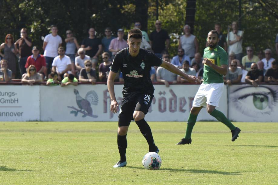 Ein Fußballspieler in einem schwarzen Trikot dribbelt den Ball auf dem Spielfeld, während eine Menge Zuschauer vom Spielfeldrand aus zusieht.
