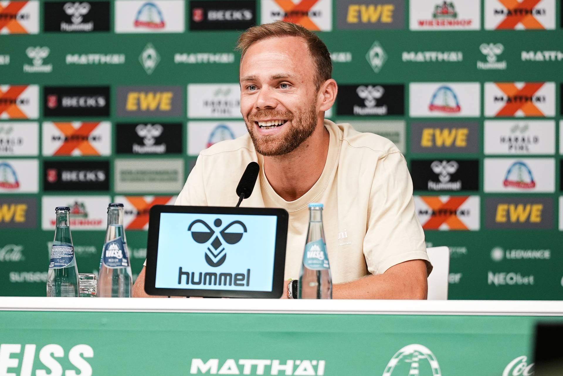 Ein lächelnder Mann spricht auf einer Pressekonferenz, während er vor einem Mikrofon und einer Kulisse mit verschiedenen Logos sitzt.