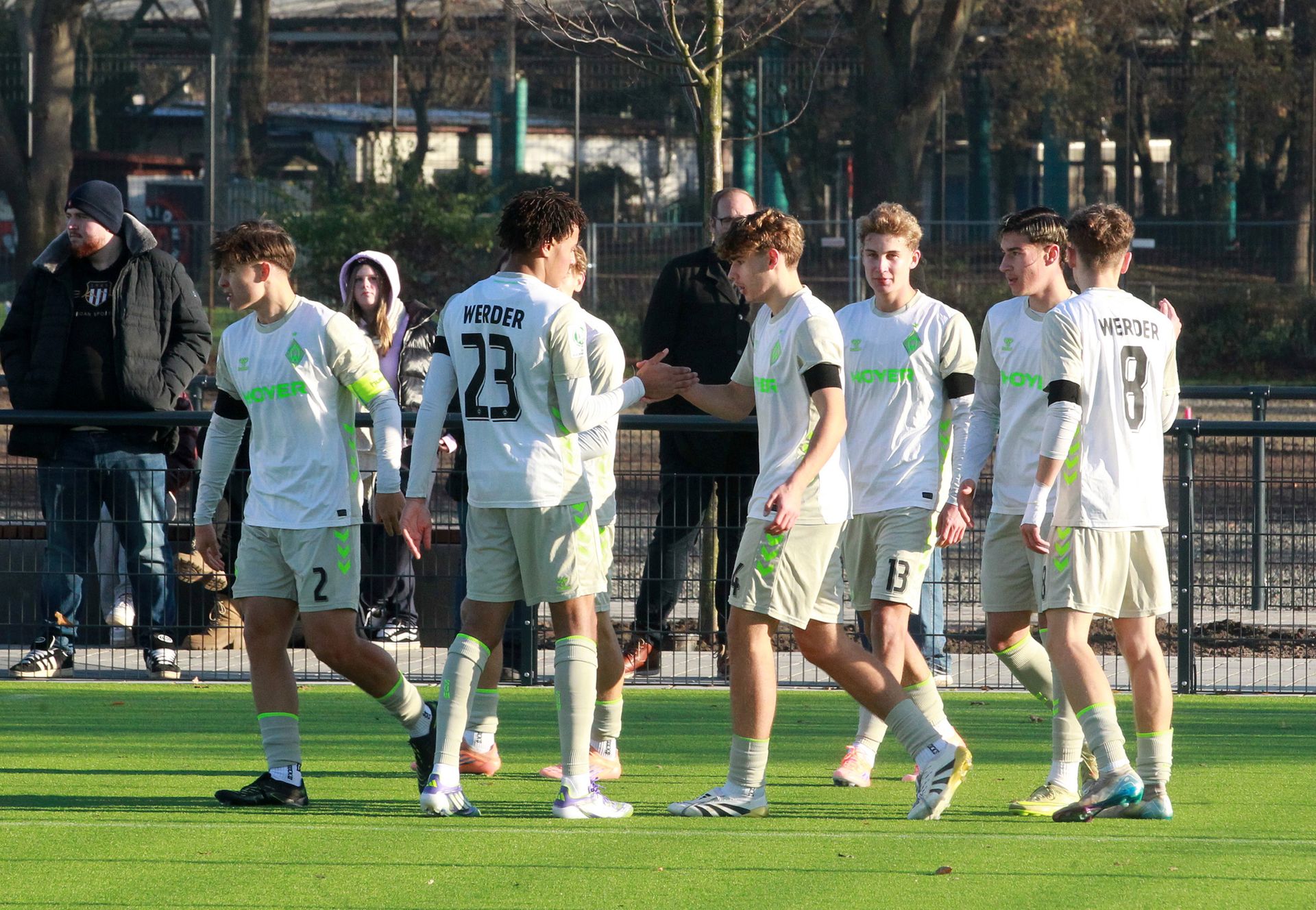 Junioren-Spieler von Werder in weiß-grünen Trikots klatschen sich ab, im Hintergrund Zuschauer und Bäume.