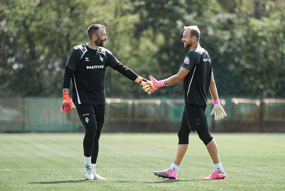 Zwei Fußballspieler in Trainingskleidung geben sich auf einem Rasenfeld ein High-Five. Beide tragen Handschuhe und lächeln, im Hintergrund sind Bäume zu sehen.