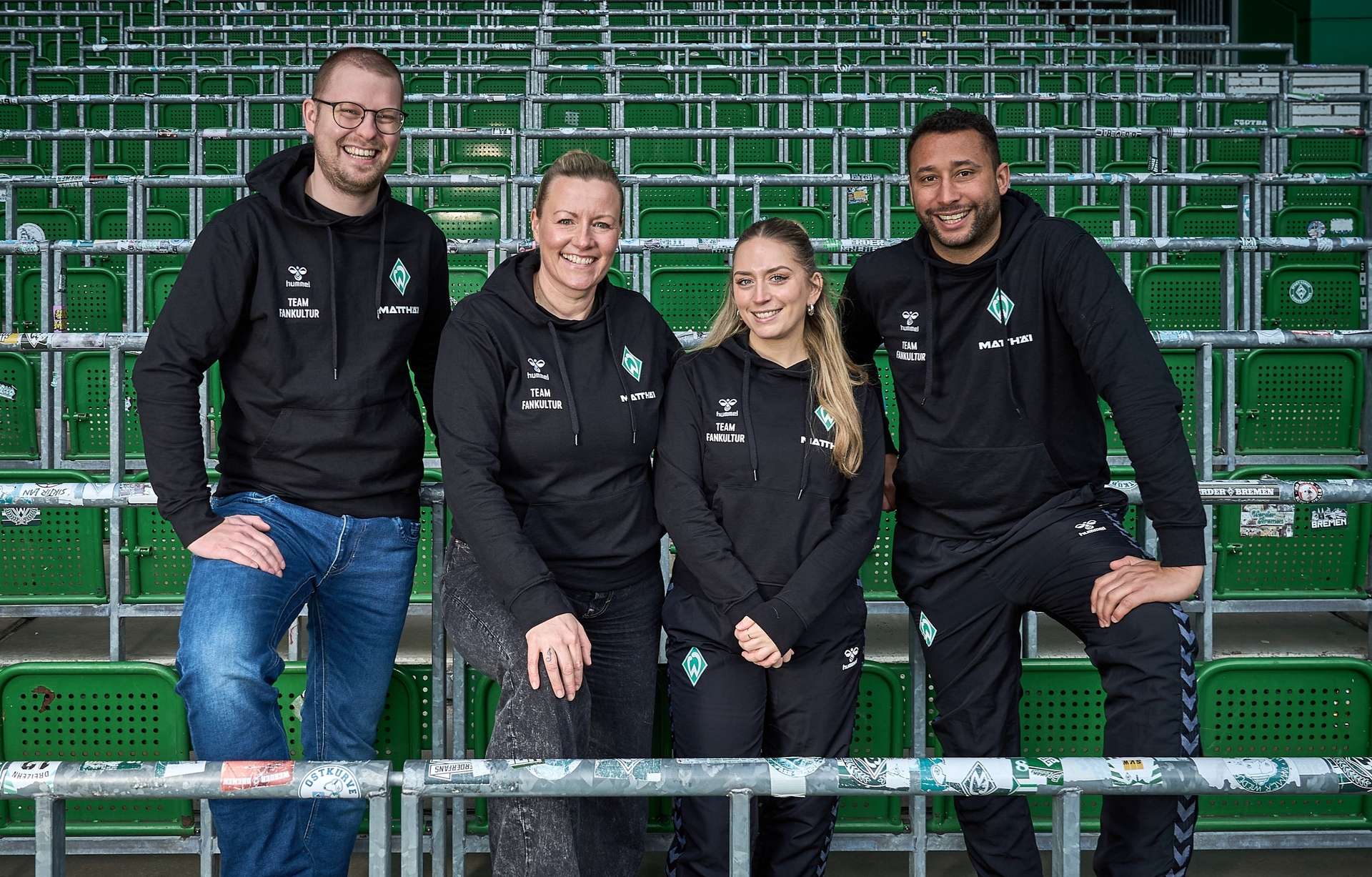 Vier Personen in identischen schwarzen Kapuzenpullis stehen und sitzen auf grünen Stadion-Sitzen und lächeln in die Kamera.