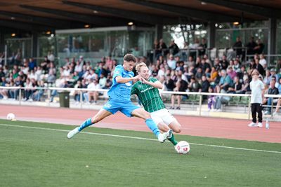 Ein Spieler der U23 von Werder Bremen streckt seinen Fuß nach dem Ball aus, er trägt ein grünes Trikot. Der Gegner im hellblauen Trikot versucht ebenfalls mit dem ausgestreckten Bein noch den Ball zu erreichen, während im Hintergrund Zuschauer von den Tribünen aus zuschauen.