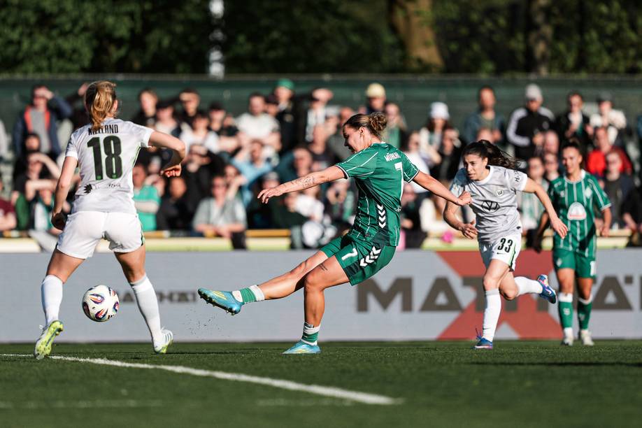 Larissa Mühlhaus im grünen Trikot schießt, eine Wolfsburger Spielerin im weißen Trikot mit der Nummer 18 auf dem Rücken steht daneben.