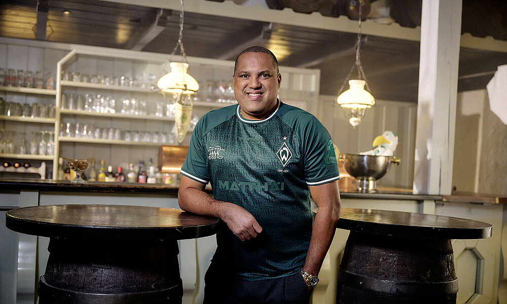 Ailton trägt das Jubiläumstrikot. Er lehnt an einem Fass