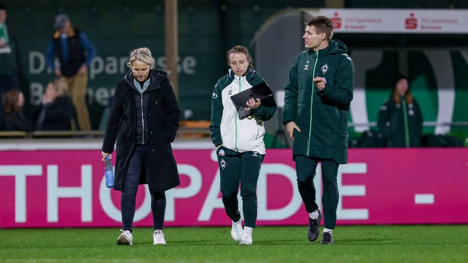 Fritzy Kromp, Michaela Specht und Björn Bremermann laufen über den Rasen und sprechen miteinander. Im Hintergrund ist eine pinke Werderbande zu sehen.