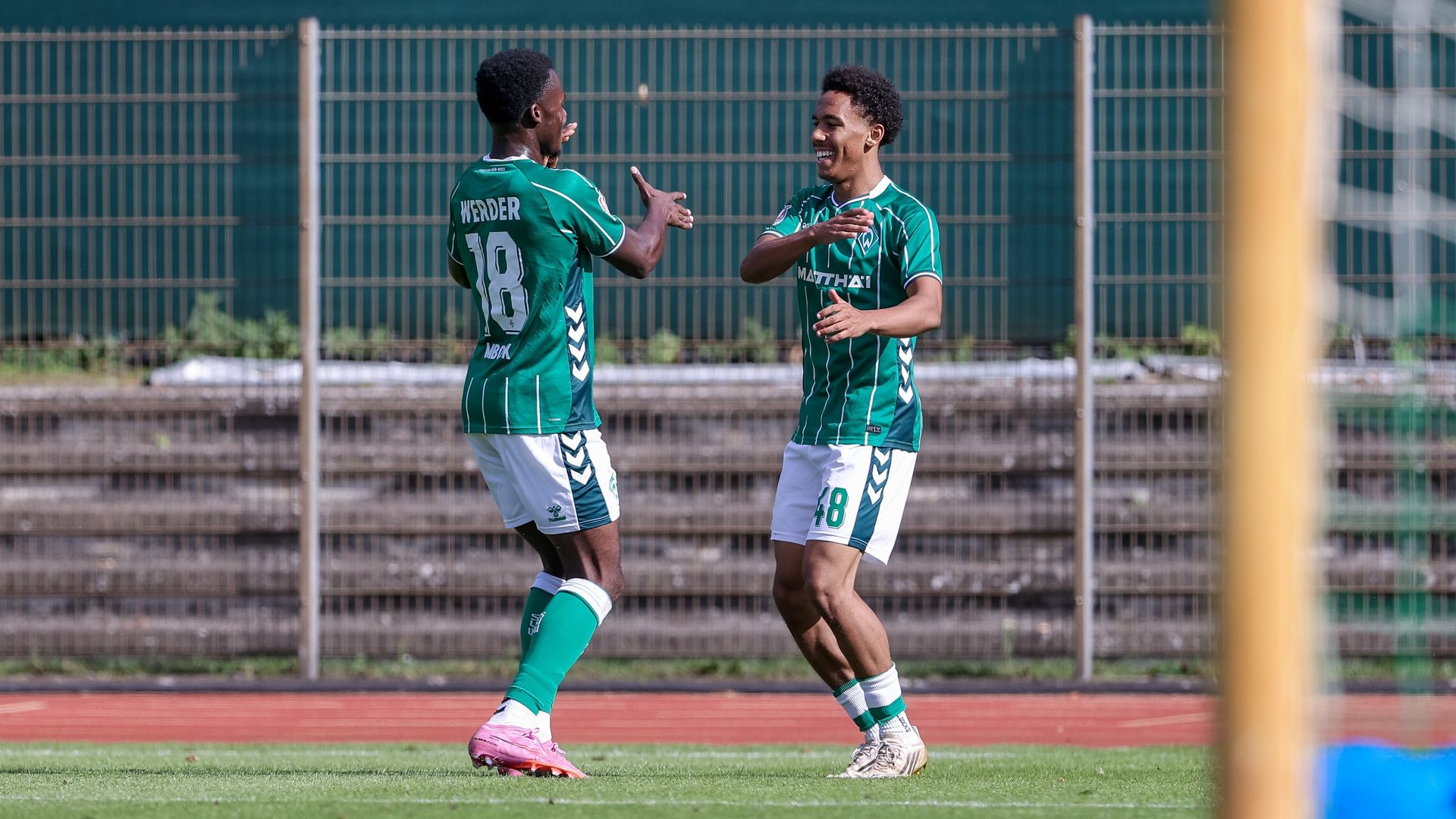 Zwei Fußballspieler in grünen Trikots feiern mit einem Lächeln und High Fives auf dem Spielfeld, vor dem Hintergrund eines Torpfostens und eines Drahtzauns.