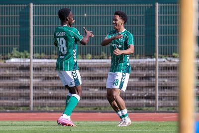 Zwei Fußballspieler in grünen Trikots feiern mit einem Lächeln und High Fives auf dem Spielfeld, vor dem Hintergrund eines Torpfostens und eines Drahtzauns.