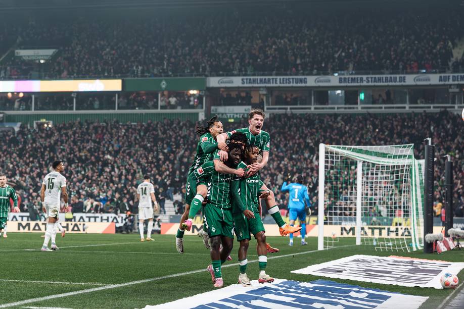 Fußballspieler in grünen Trikots feiern ein Tor, indem sie sich in der Nähe der Eckfahne auf einen Mitspieler stürzen, während das vollbesetzte Stadion zusieht.
