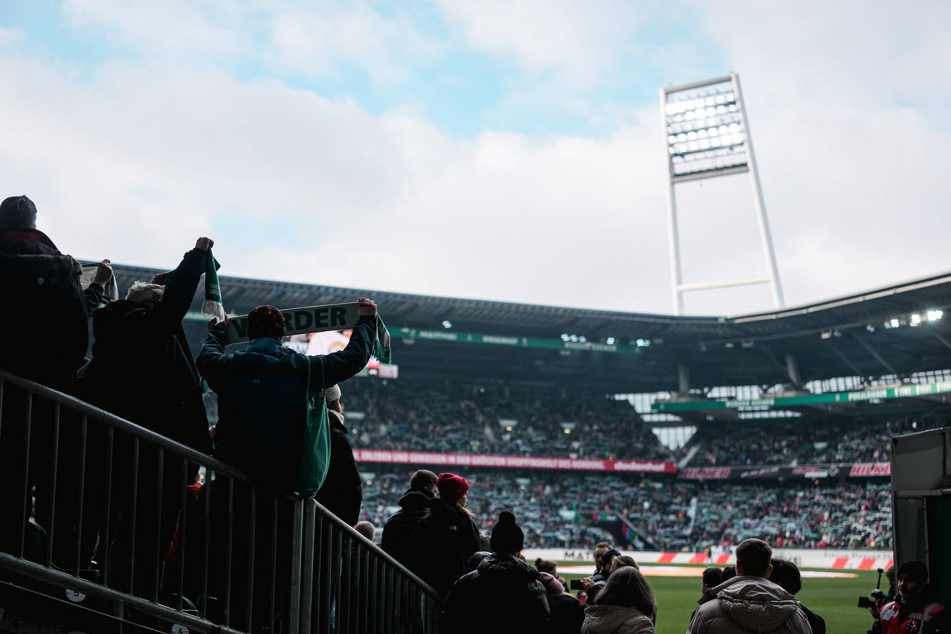 Fans jubeln im Weserstadion, mit Blick auf das Spielfeld und einer großen Menschenmenge unter bewölktem Himmel.