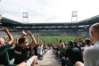 Die Fans jubeln in einem vollbesetzten Fußballstadion und recken vor Begeisterung die Hände in die Höhe, während sich das Spiel auf dem grünen Rasen unter ihnen entfaltet.