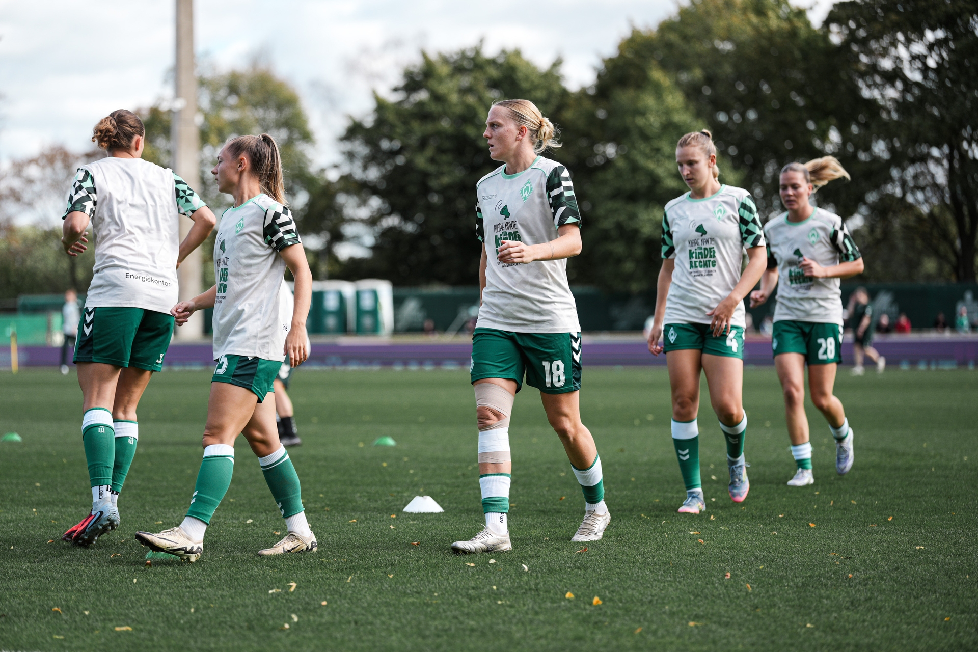 Soccer players in green and white uniforms warming up on a grassy field, with trees in the background.