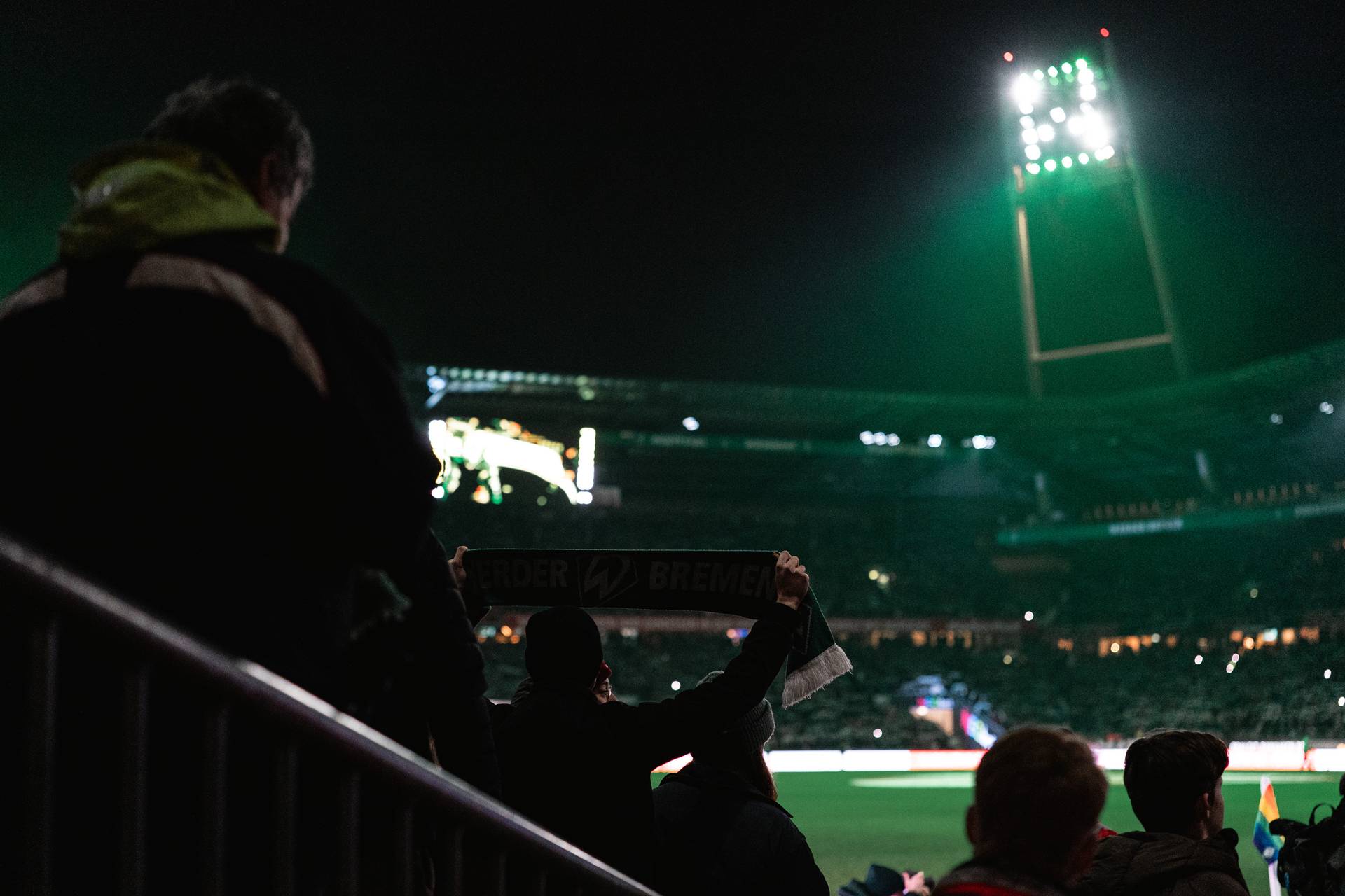 Fans halten einen Werder-Bremen-Schal in einem schwach beleuchteten Stadion bei Nacht, während helle Flutlichter das Spielfeld erhellen.