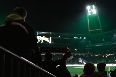 Fans halten einen Werder-Bremen-Schal in einem schwach beleuchteten Stadion bei Nacht, während helle Flutlichter das Spielfeld erhellen.