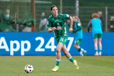 Lara Schmidt in einem grünen Trikot dribbelt den Ball auf dem Spielfeld, im Hintergrund sind ihre Teamkolleginnen und eine blaue Bandenwerbung zu sehen.