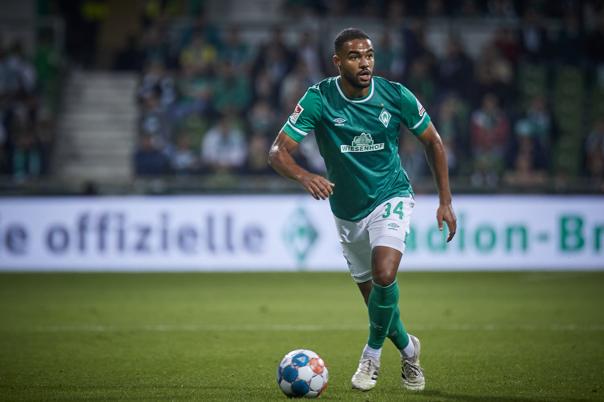 Ein Fußballspieler im grünen Trikot mit der Nummer 34 dribbelt während eines Spiels auf dem Platz den Ball, im Hintergrund ist eine verschwommene Zuschauermenge zu sehen.