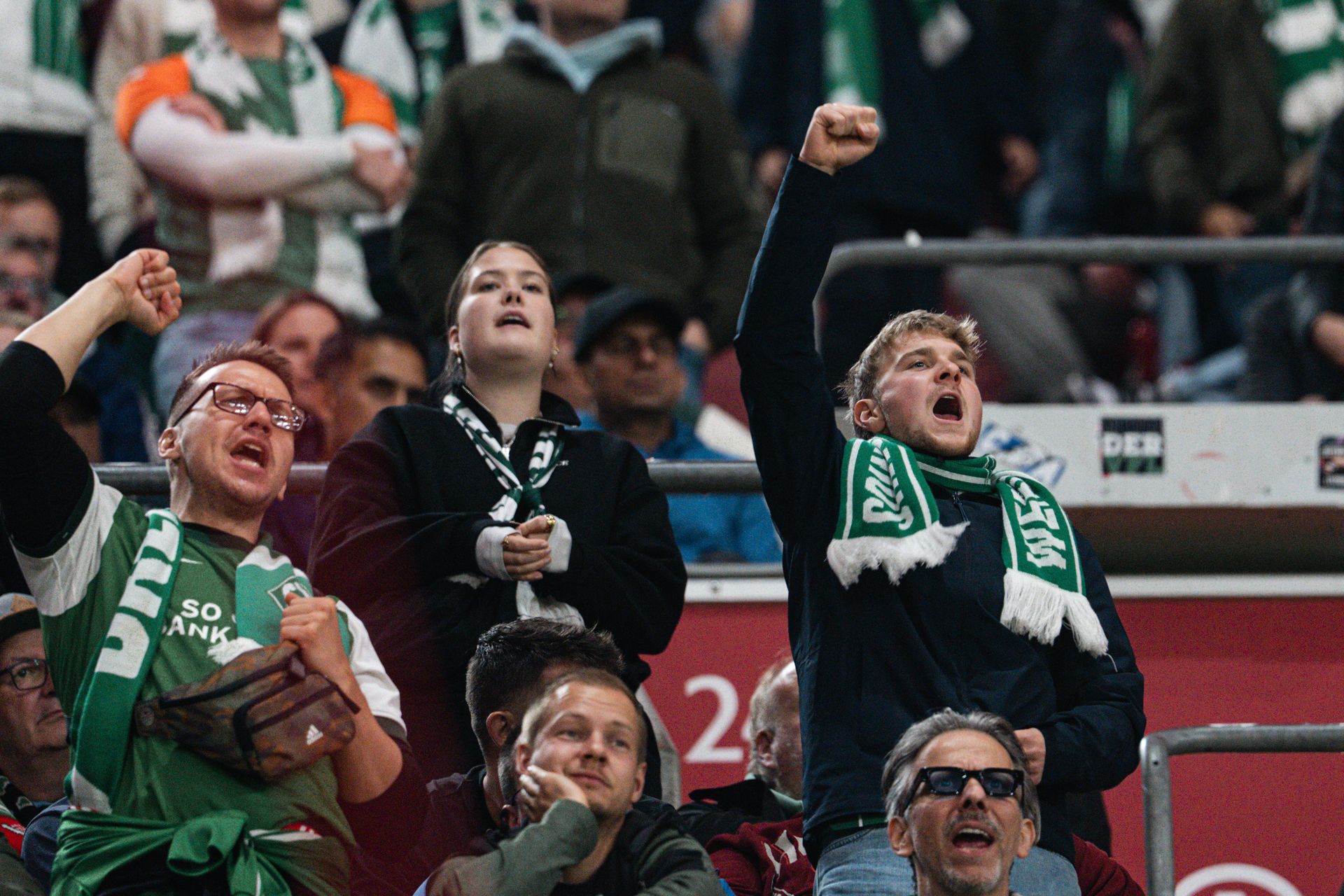 Werder-Fans mit Schal stehen auf in München auf der Tribüne