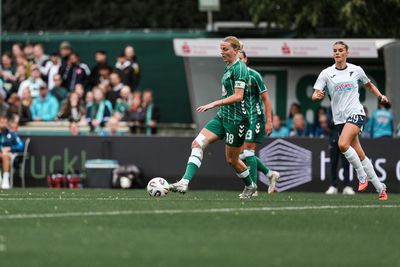 Lina Hausicke in einer grünen Uniform dribbelt den Ball auf einem Feld, verfolgt von einer Gegnerin in einer weißen Uniform. Im Hintergrund stehen Zuschauer.