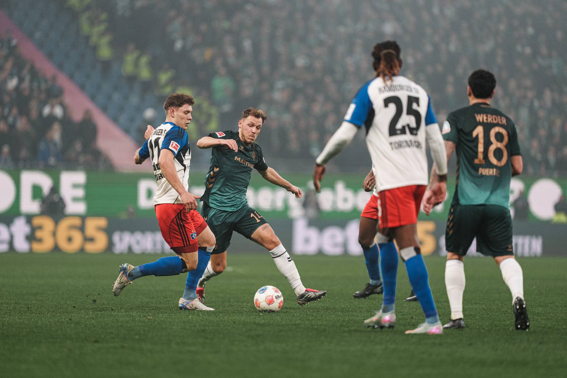 Fußballspieler in Aktion während eines Spiels auf einem Rasenplatz, umgeben von einer großen Zuschauermenge und Stadionwerbung.