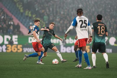 Fußballspieler in Aktion während eines Spiels auf einem Rasenplatz, umgeben von einer großen Zuschauermenge und Stadionwerbung.