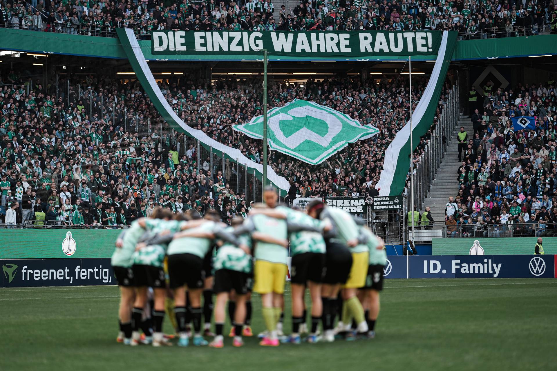 Die WERDERFRAUEN stehen im Kreis, im Hintergrund ist die Fankurve von Werder zu sehen, die eine Choreo hochhalten.