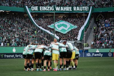 Die WERDERFRAUEN stehen im Kreis, im Hintergrund ist die Fankurve von Werder zu sehen, die eine Choreo hochhalten.
