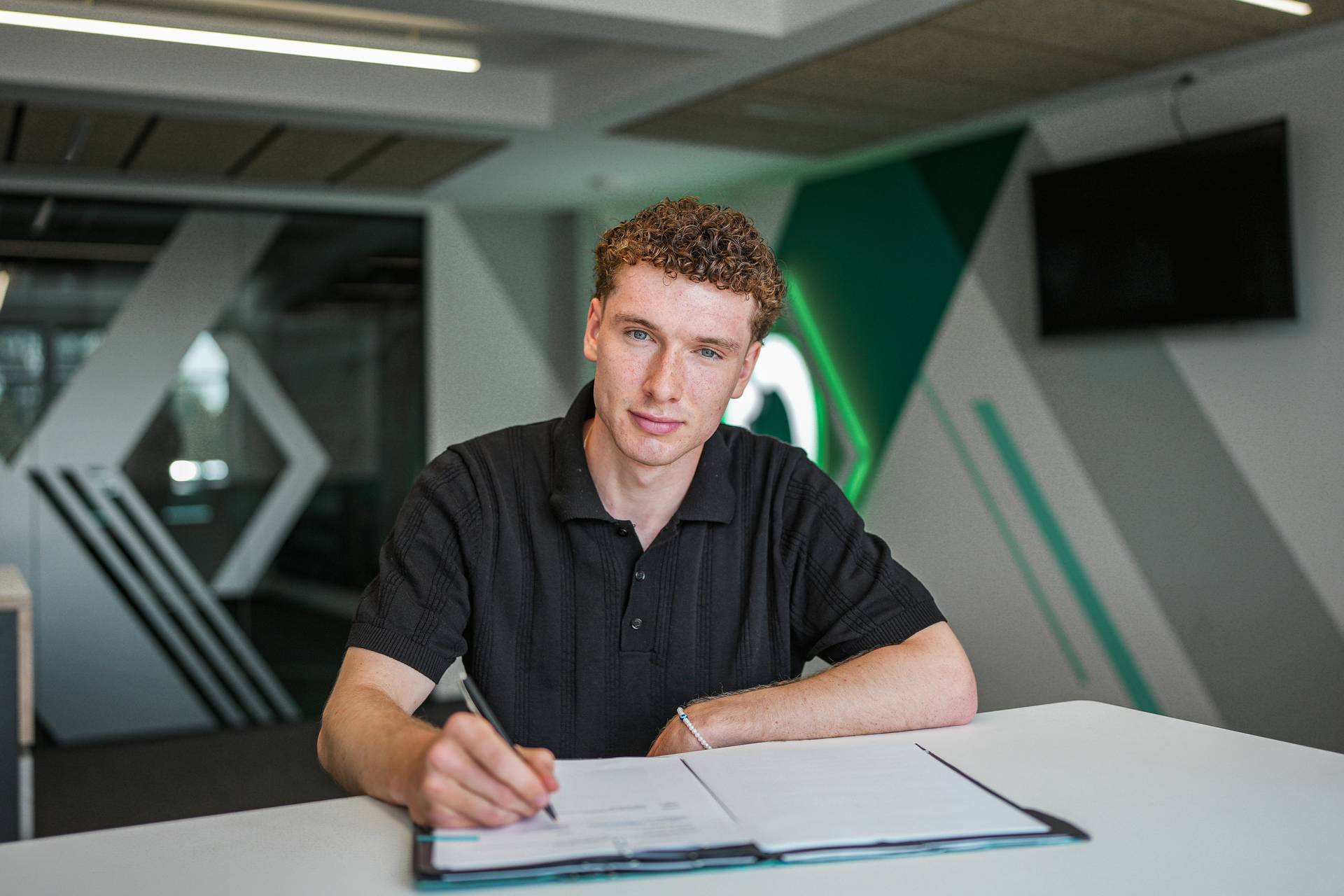 Patrick Götzelmann sitzt an einem Schreibtisch, hält einen Stift in der Hand und hat eine Mappe mit Papierblättern vor sich liegen.