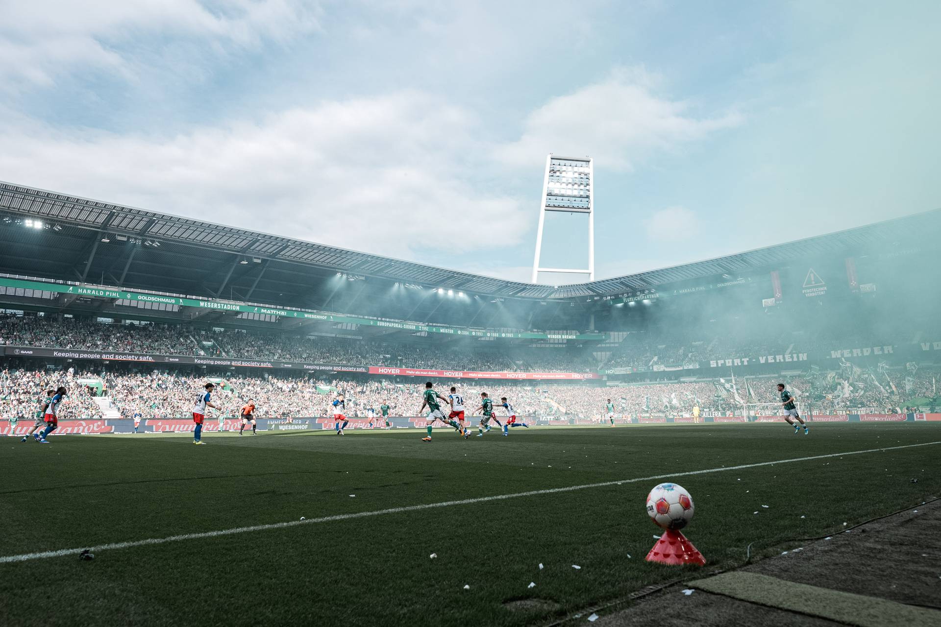 Ein Fußballspiel in einem vollbesetzten Stadion, in dem die Spieler über den Rasen laufen und eine bunte Rauchwolke über ihnen schwebt, unter einem blauen, bewölkten Himmel.