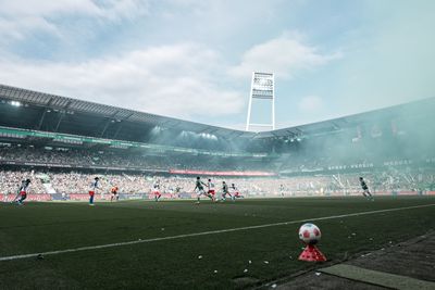 Ein Fußballspiel in einem vollbesetzten Stadion, in dem die Spieler über den Rasen laufen und eine bunte Rauchwolke über ihnen schwebt, unter einem blauen, bewölkten Himmel.