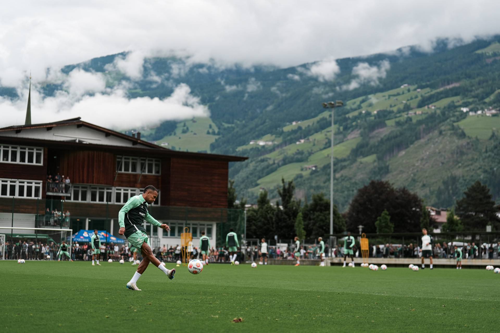 Ein Fußballspieler in grüner Kleidung schießt einen Ball auf einem üppigen Rasen, im Hintergrund sind Berge und ein bewölkter Himmel zu sehen.