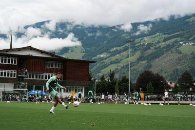 Ein Fußballspieler in grüner Kleidung schießt einen Ball auf einem üppigen Rasen, im Hintergrund sind Berge und ein bewölkter Himmel zu sehen.
