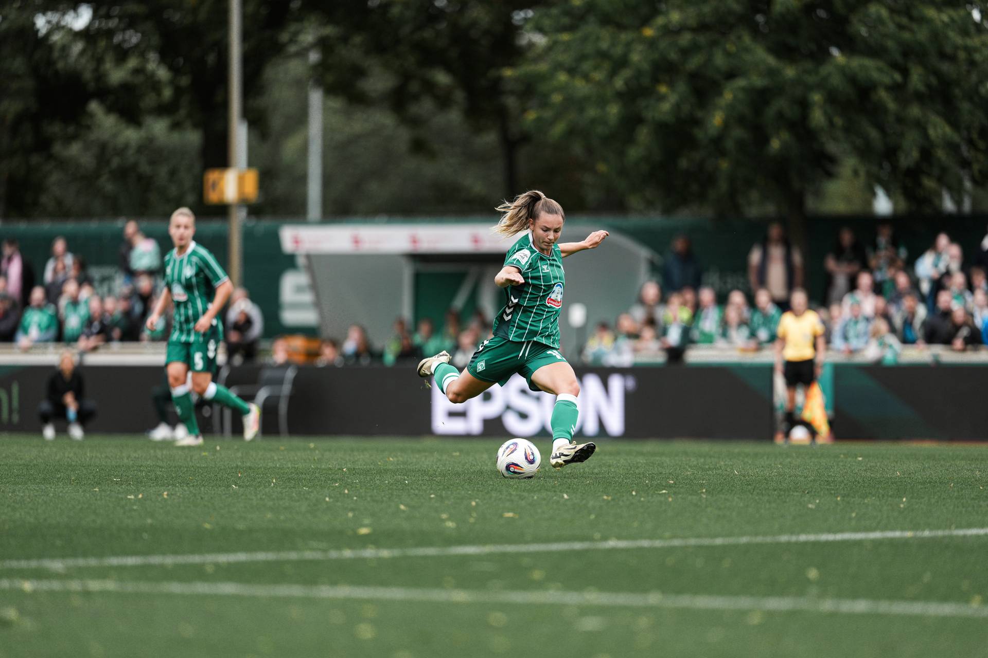 Michelle Weiß am Ball auf Platz 11. Sie setzt zum Schuss an. Im Hintergrund sieht man Fans und die Trainer*innenbank.
