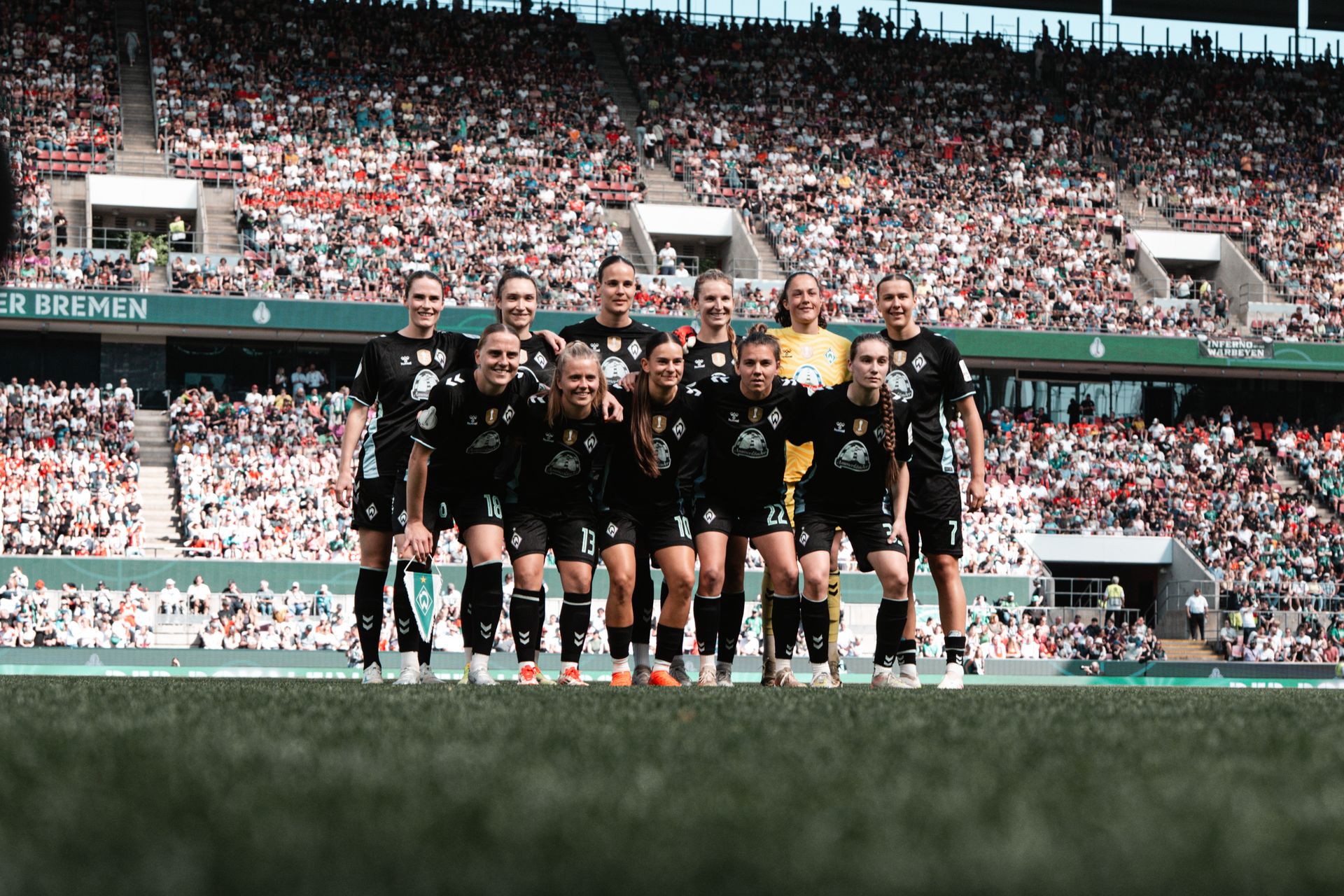 Die WERDERFRAUEN in schwarzen Trikots posiert auf dem Spielfeld in einem Stadion voller Zuschauer.
