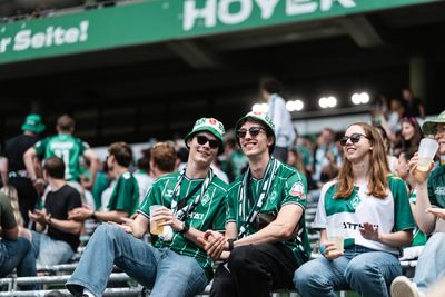 Drei Werder-Fans mit Hüten, Schaals und Sonnenbrillen sitzen gut gelaunt in der Osrkurve auf den Wellenbrechern.