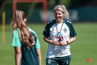 Fritzy Kromp lächelnd auf dem Trainingsplatz im weißen Oberteil. Im Vordergrund ist unscharf eine Spielerin zu sehen, die Kromp anschaut.