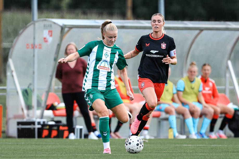 Eine Spielerin im grün-weiß gestreiftem Fußballtrikot spielt einen Pass, neben ihr läuft eine Spielerin im schwarzen Trikot und roter Sporthose.