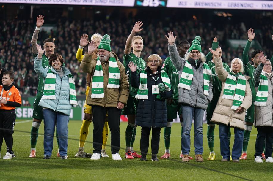 Eine Gruppe von Menschen auf einem Fußballplatz, die grün-weiße Schals und Wintermäntel tragen, lächeln und der Menge zuwinken.