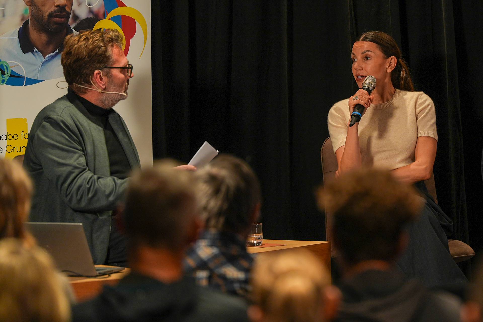 Ein Mann und eine Frau sitzen während einer Podiumsdiskussion auf der Bühne. Die Frau hält ein Mikrofon, der Mann einen Zettel in der Hand. Im Vordergrund sind Zuschauer zu sehen.