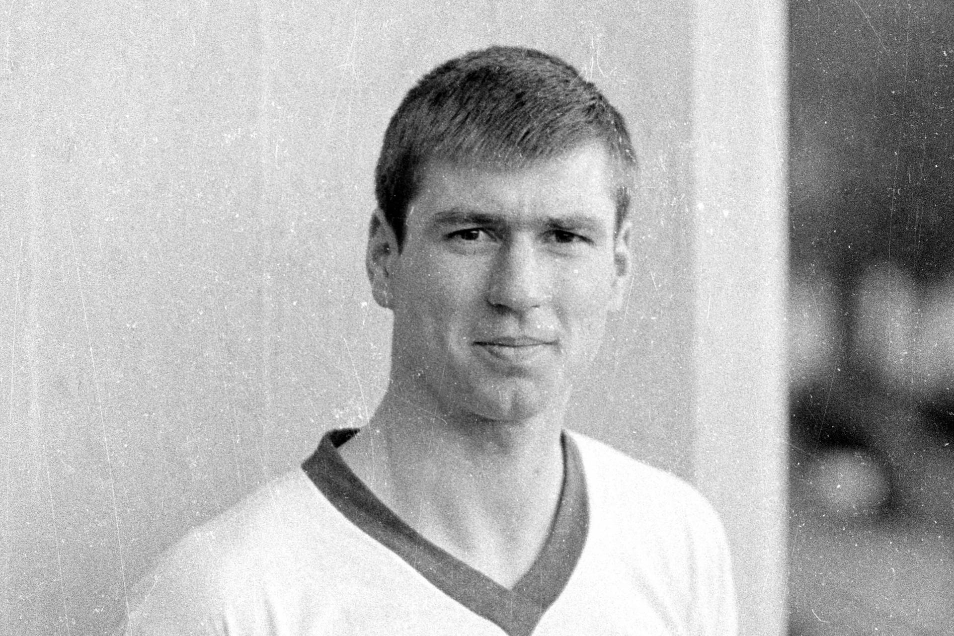 Schwarz-weiß Bild von Sepp Piontek. Er steht vor einer Wand und trägt ein weißes Shirt mit dunklem Kragen.