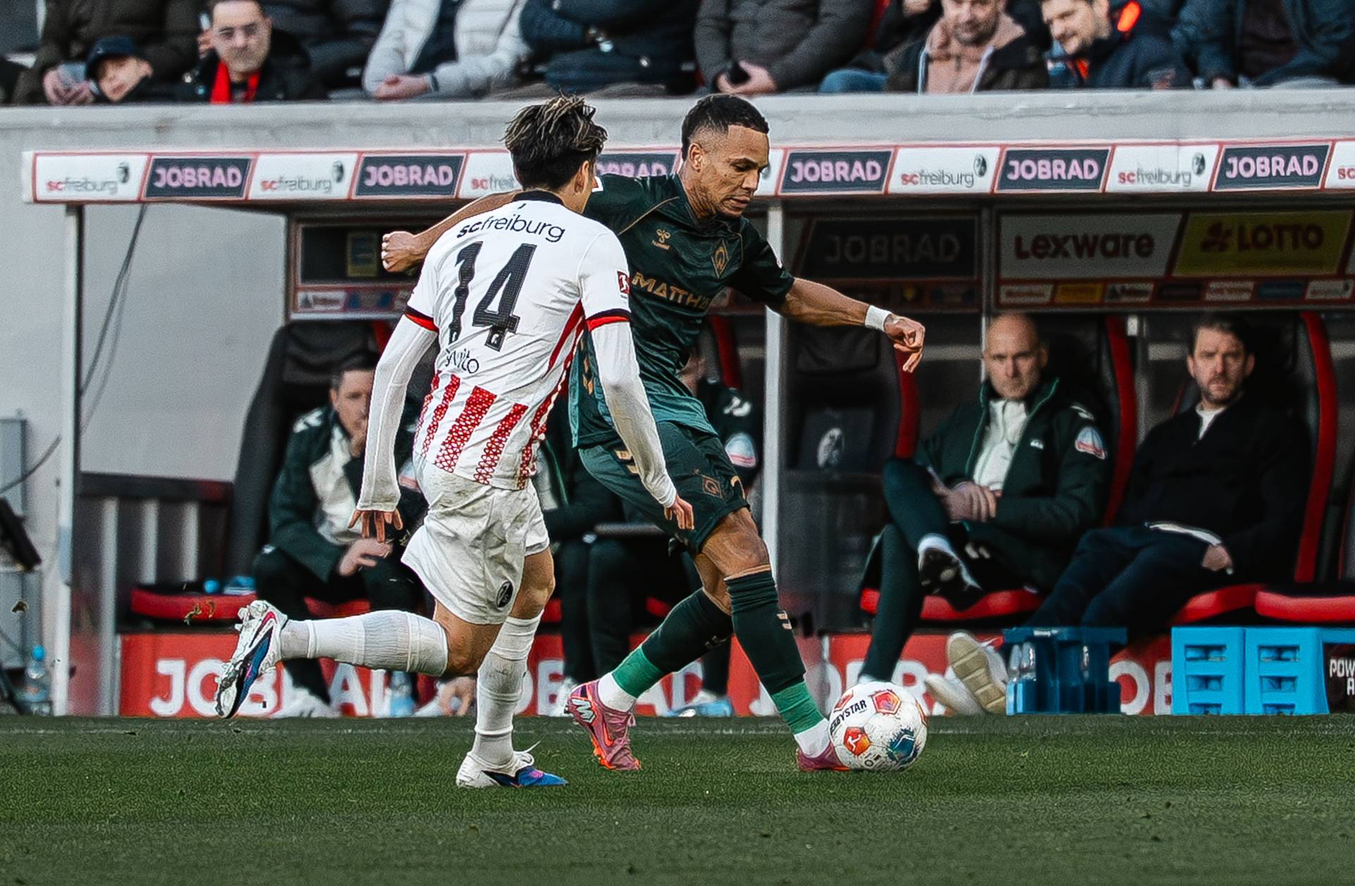 Felix Agu läuft mit dem Ball am Fuß. Ein Freiburger Spieler läuft auf ihm zu. Im Hintergrund ist die Trainerbank zu sehen.