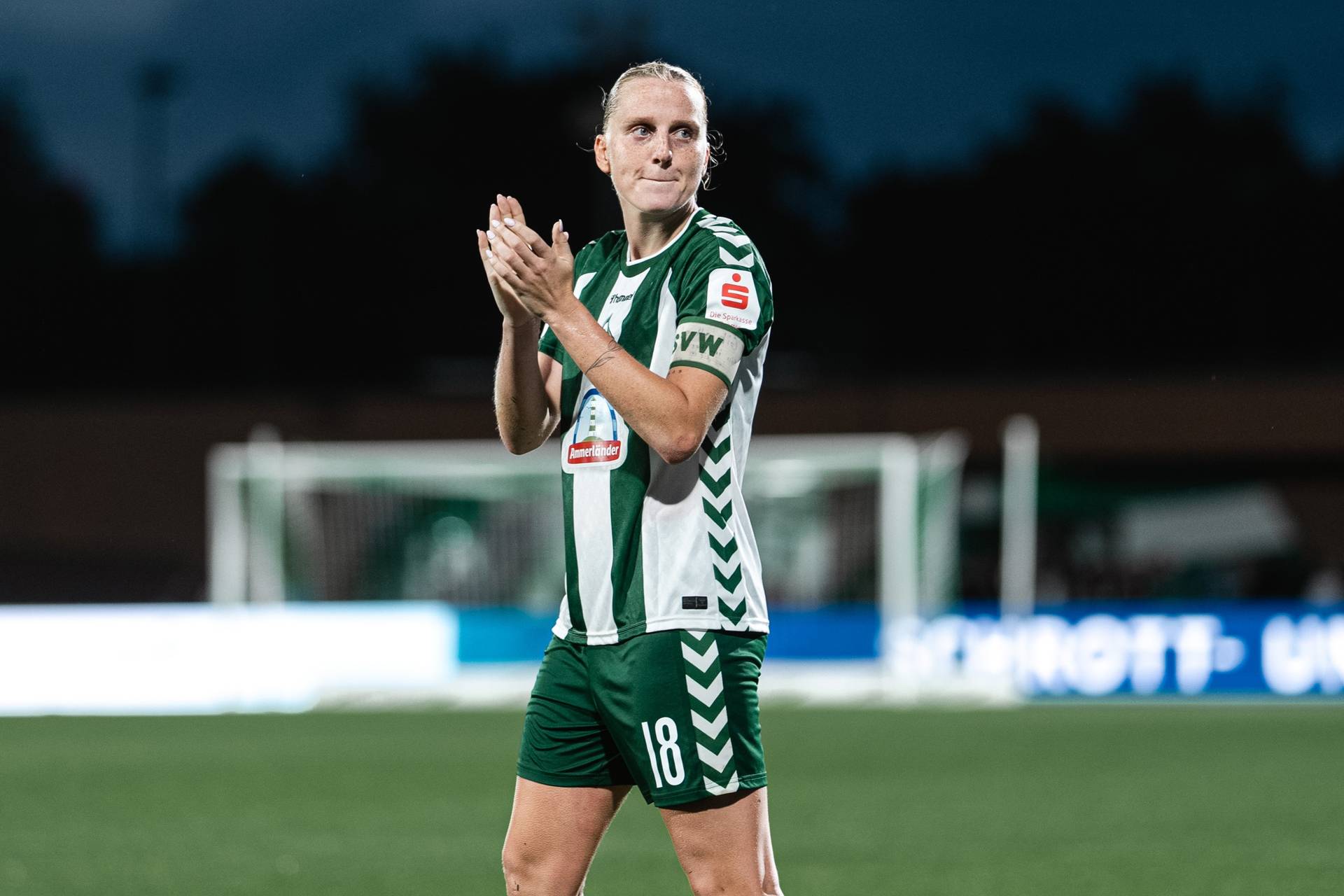 Ein Fußballspieler in einem grün-weißen Trikot klatscht auf dem Spielfeld, im Hintergrund sind ein verschwommener Torpfosten und Stadionlichter zu sehen.