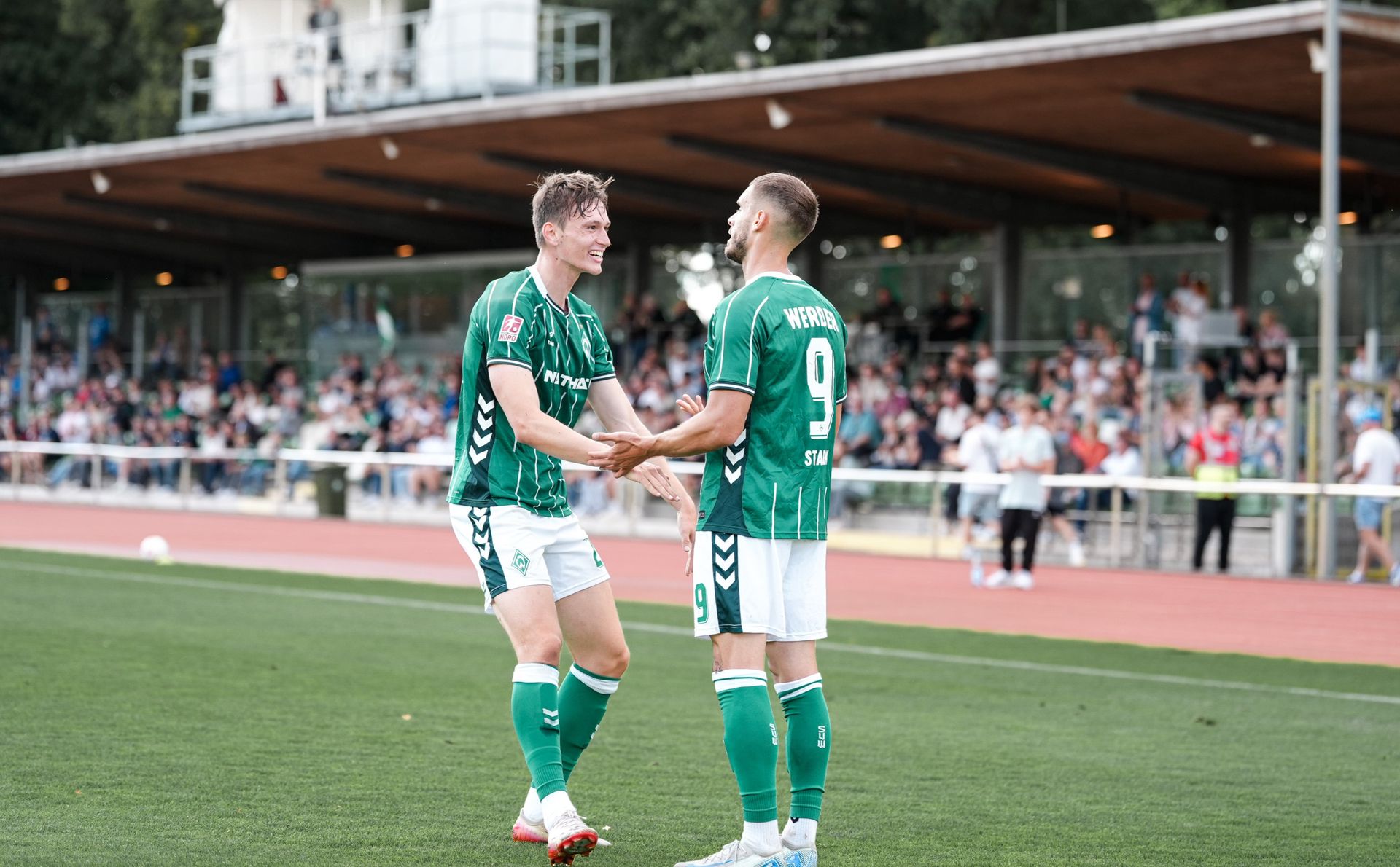 Im Zwei Spieler der U23 des SVW (in grünen Trikots) klatschen ab und freuen sich. Hintergrund sieht eine Menschenmenge von der Tribüne aus zu.
