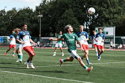 Fußballspieler in grünen und blauen Trikots kämpfen während eines Spiels an einem sonnigen Tag vor dem Hintergrund von Bäumen in der Luft um den Ball.