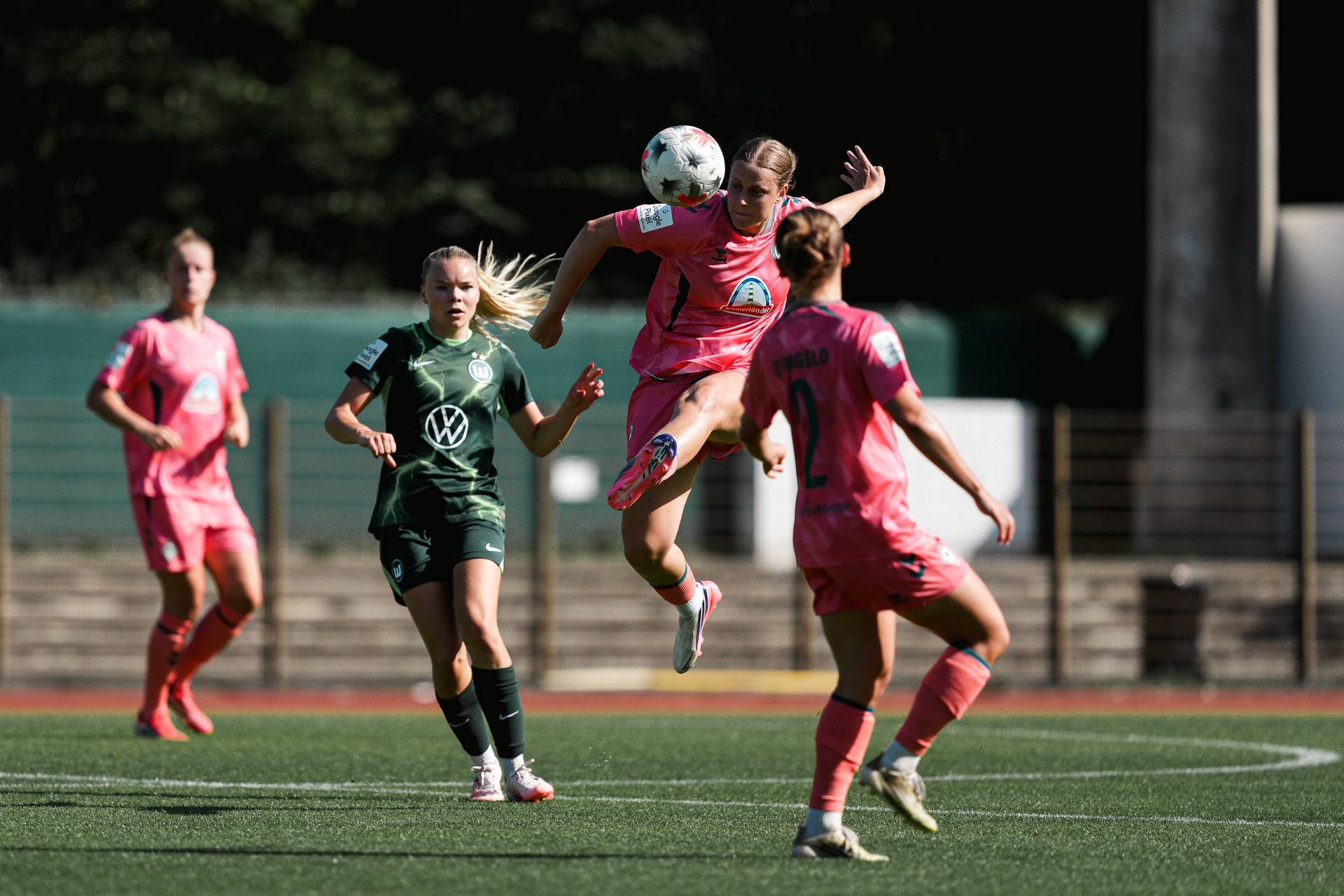Ein Fußballspieler in einem rosa Trikot führt einen Sprungschuss aus, umgeben von Mannschaftskameraden und Gegnern auf einem grünen Rasenfeld.
