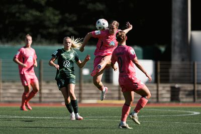 Ein Fußballspieler in einem rosa Trikot führt einen Sprungschuss aus, umgeben von Mannschaftskameraden und Gegnern auf einem grünen Rasenfeld.