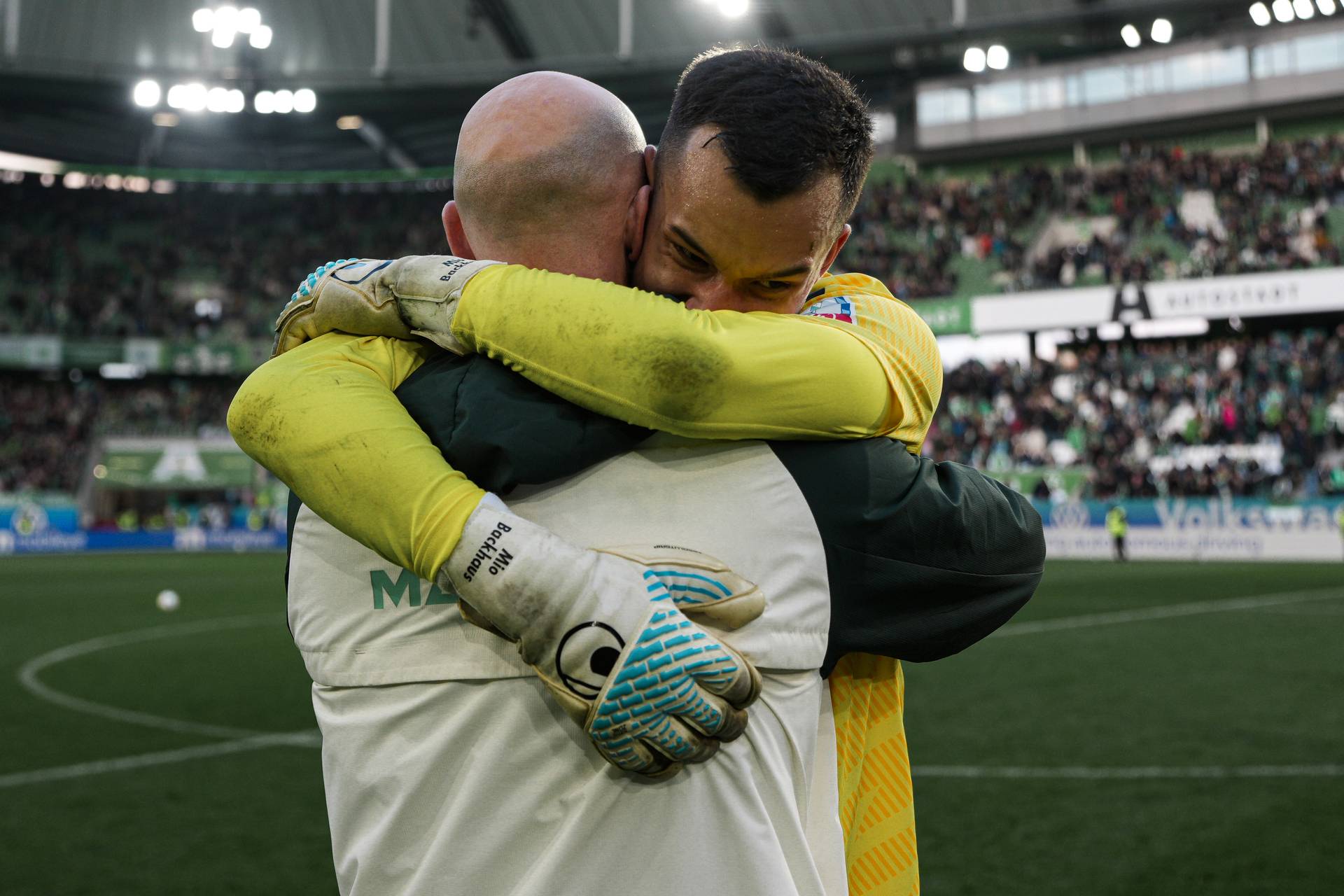 Mio Backhaus im gelben Trikot umarmt einen Mann mit weißer Jacke und Glatze. Im Hintergrund ist das Stadion des VfL Wolfsburg zu sehen.