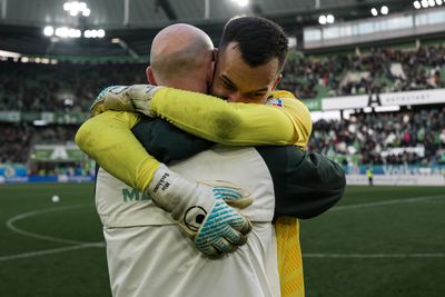 Mio Backhaus im gelben Trikot umarmt einen Mann mit weißer Jacke und Glatze. Im Hintergrund ist das Stadion des VfL Wolfsburg zu sehen.