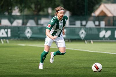 Ein Fußballspieler in einem grün-weißen Trikot rennt bei Tageslicht auf einem Rasenfeld mit einem Stirnband auf einen Fußball zu.